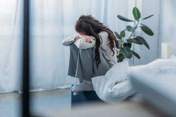 grieving woman sitting, covering face with hands and crying in bedroom