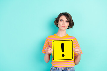 Photo of cute doubtful lady wear orange t-shirt holding exclamation point board looking empty space isolated turquoise color background