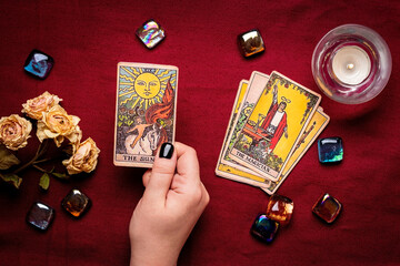 Fortuneteller's hand with black manicure lays out tarot cards, crystal, candle, dry roses on red tablecloth Flat lay Top view Fortune telling, prediction, esoteric concept